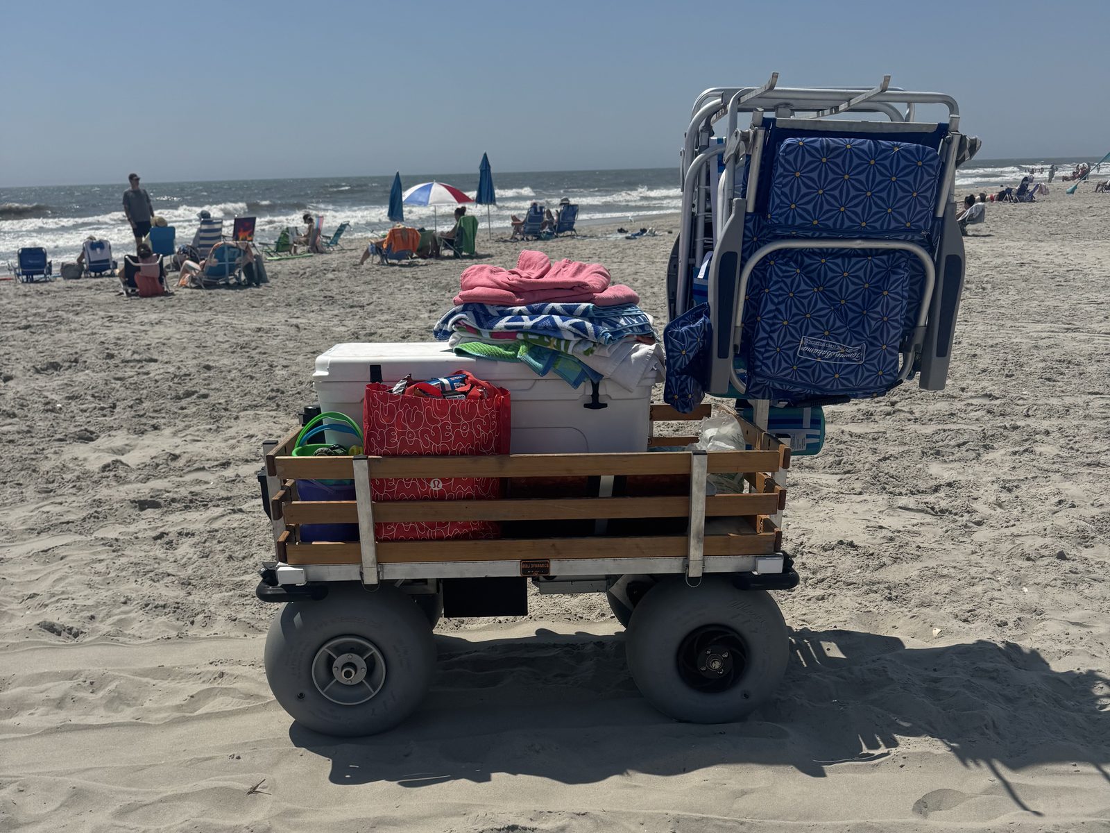 Side profile at the ocean's edge: the cart loaded with cooler, towels, and folded chairs on balloon tires